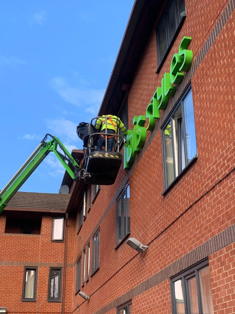 Installer on a cherry picker fitting ibis Styles 3D letters on a brick building