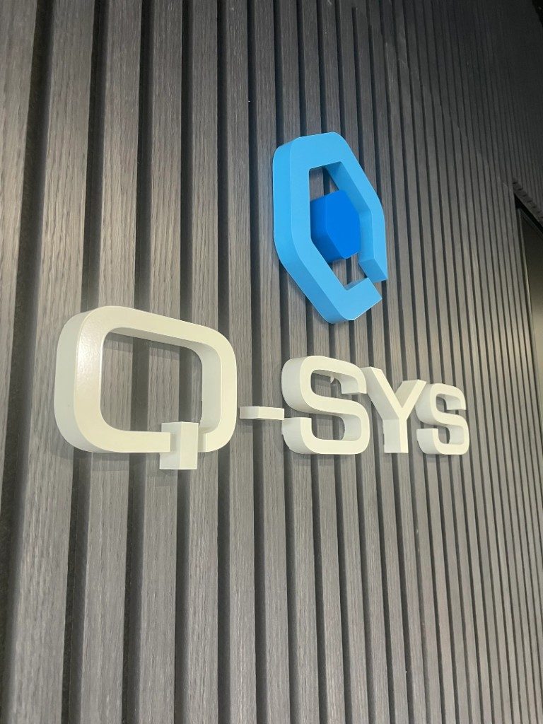 Q-SYS blue logo and white 3D lettering on a dark slatted wall